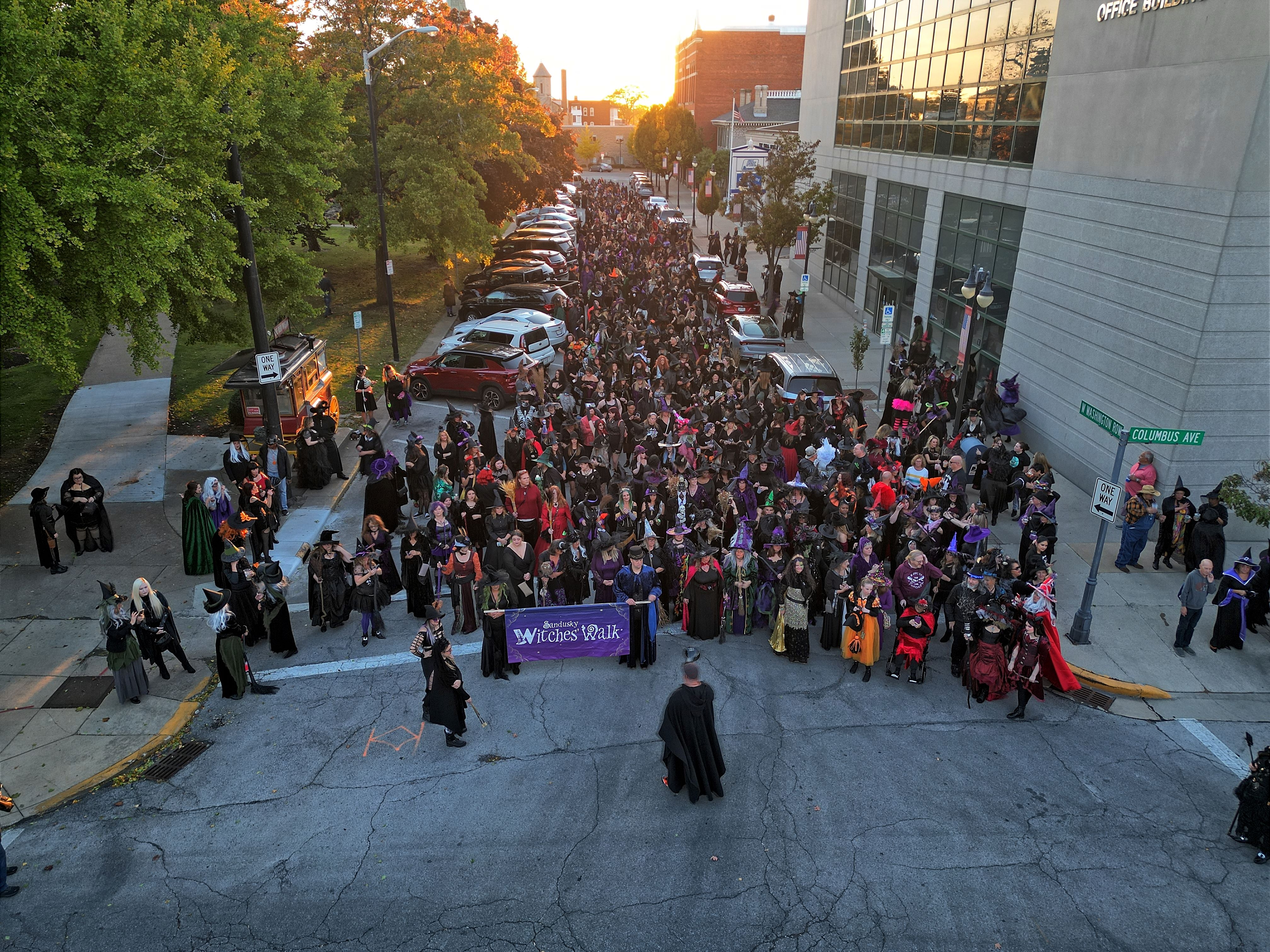 A crowd of people dressed in costumes as wicthers on a street.